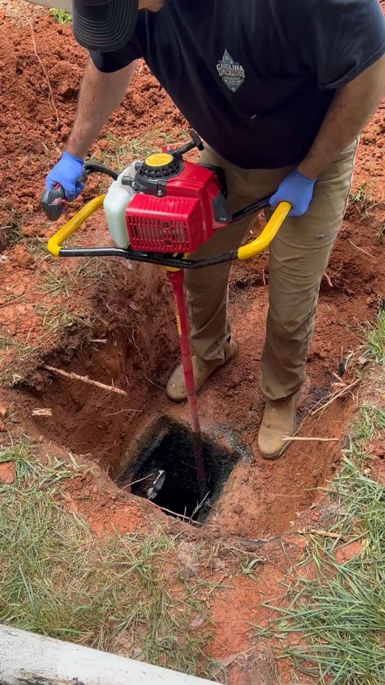 Man using a red post hole digger to drill into a square hole in red soil, outdoors.