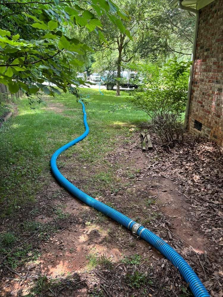 Blue hose snakes across grass, near a brick building and trees; a truck is in the distance.