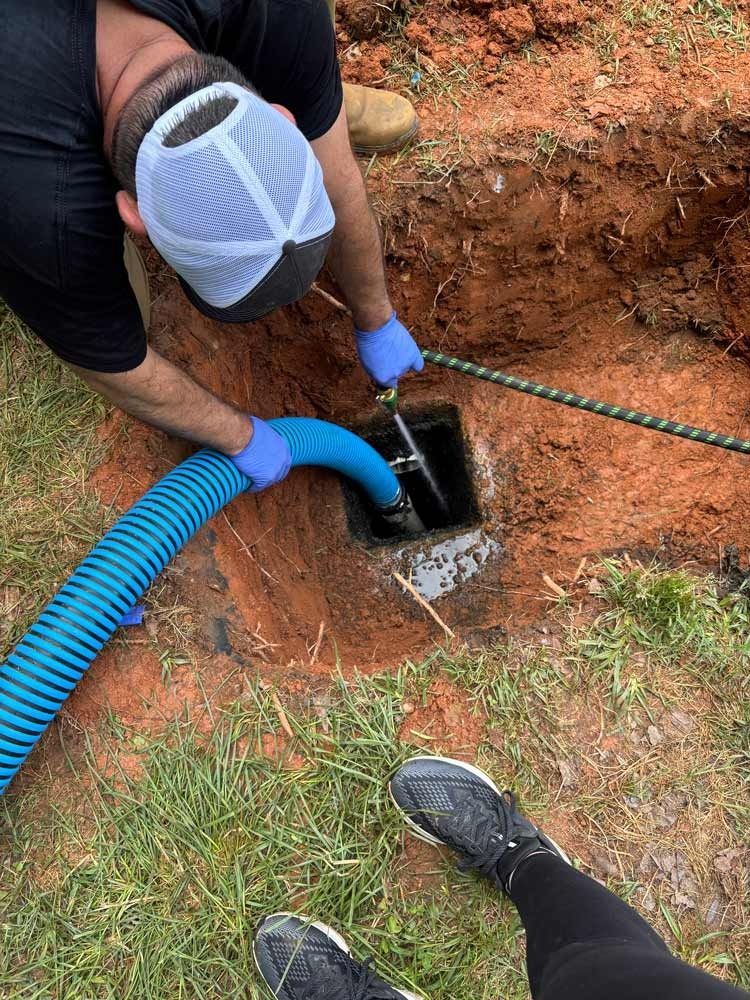 Man in blue gloves pumping septic tank. Blue hose in a square opening dug in red dirt and green grass.