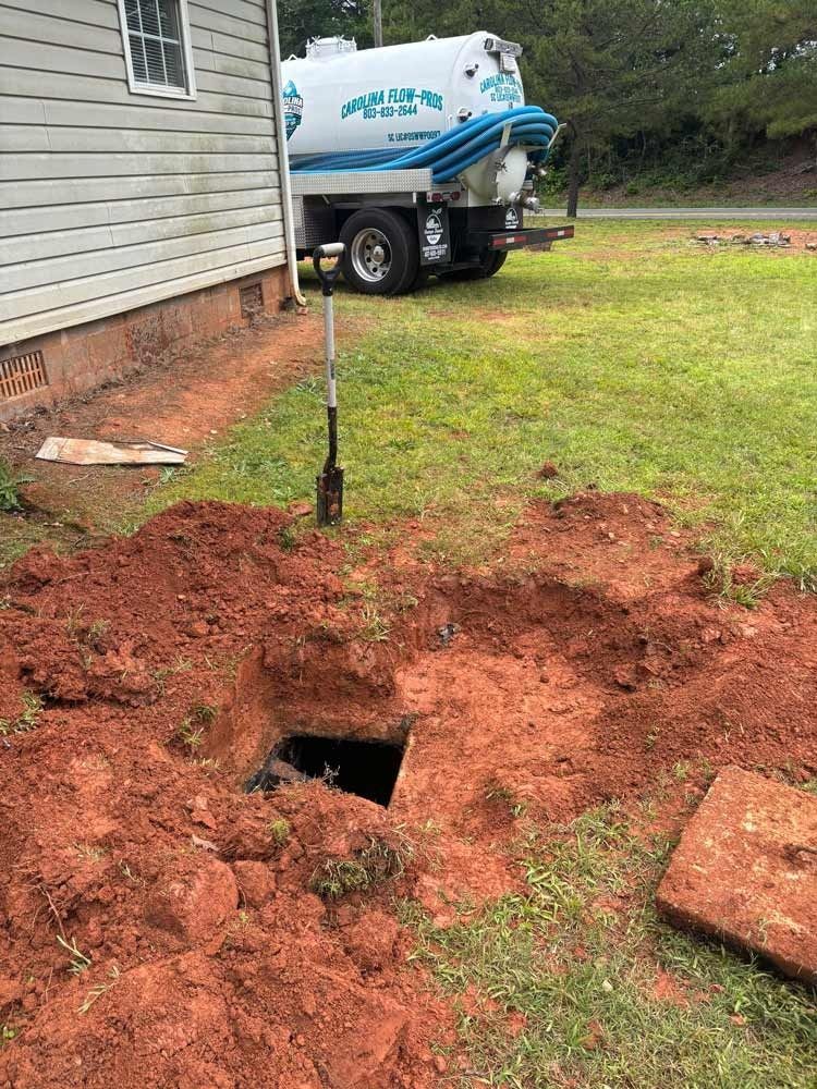 A septic tank being serviced; a truck is parked by a hole dug in red clay soil.