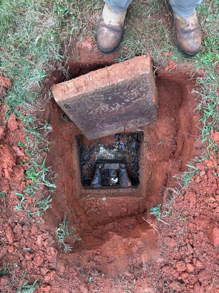 Open utility box with a removed lid in red dirt, with person's legs and boots visible.
