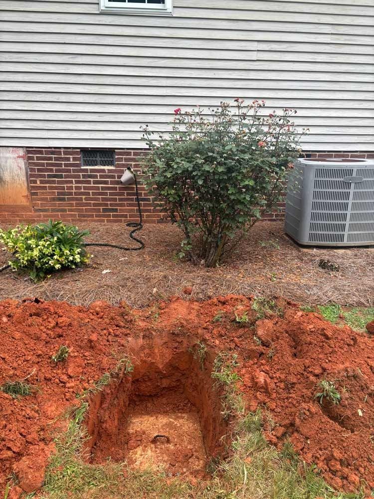 Red clay soil surrounds a square-dug hole near a brick wall, AC unit, and bush in a yard.