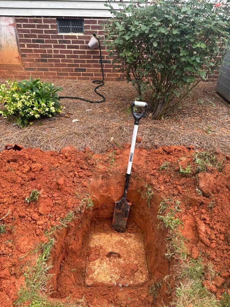 Shovel in red dirt square hole revealing concrete septic tank lid near a brick wall and bush.
