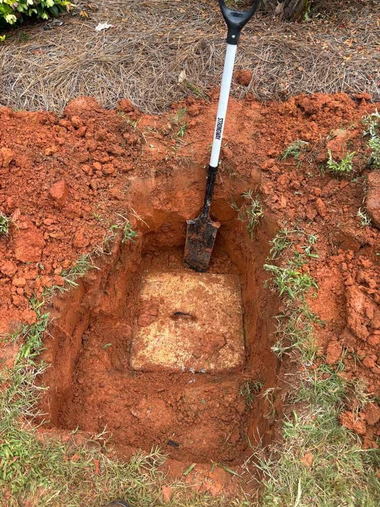 A rectangular hole in red clay soil with a shovel inside; revealing a concrete access cover.