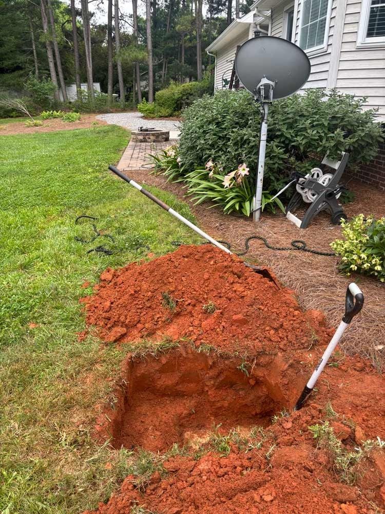 Hole dug in red clay soil in a yard, with shovels in the ground, satellite dish in background.