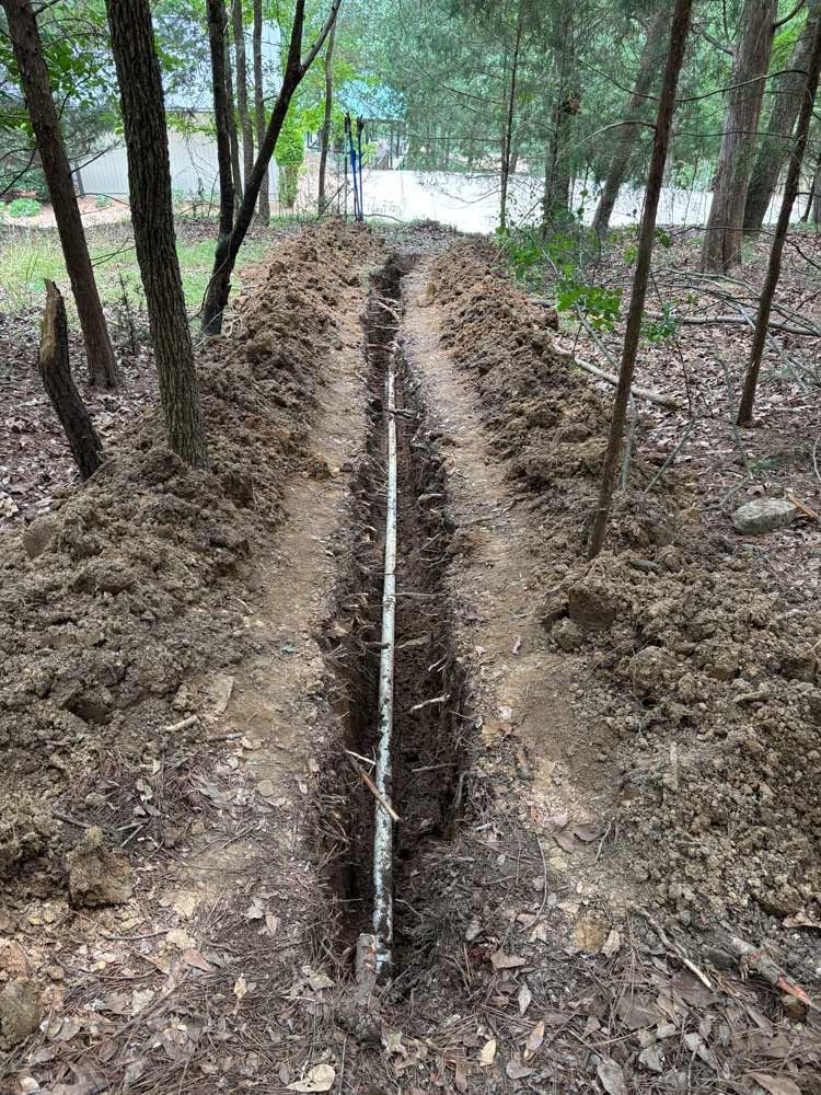 A narrow trench with a white pipe in it, dug in a wooded area.