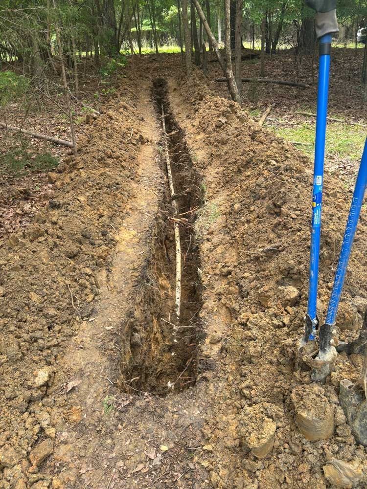 A long trench dug in the dirt with exposed pipe, two blue handled shovels are on the right.