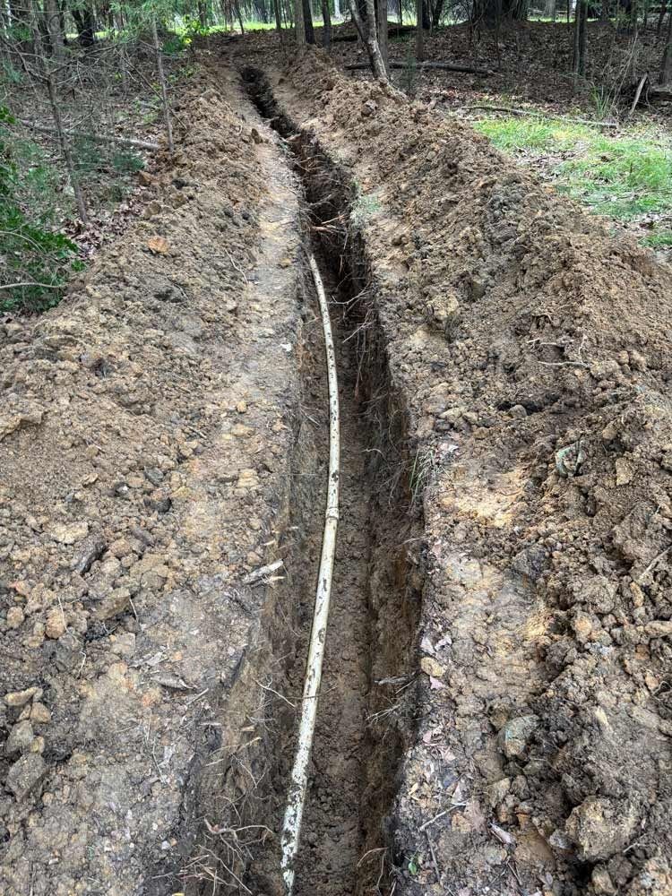 Trench dug in dirt, with a white pipe running through it, in a wooded area.