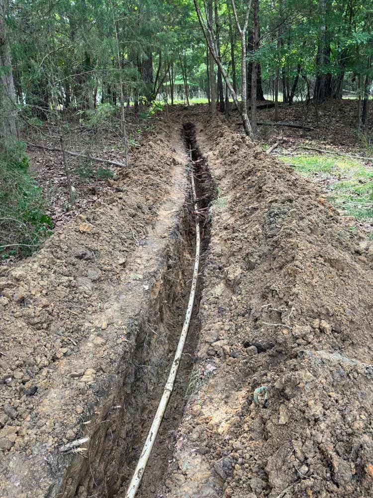A trench dug in the earth, containing white conduit, in a wooded area.