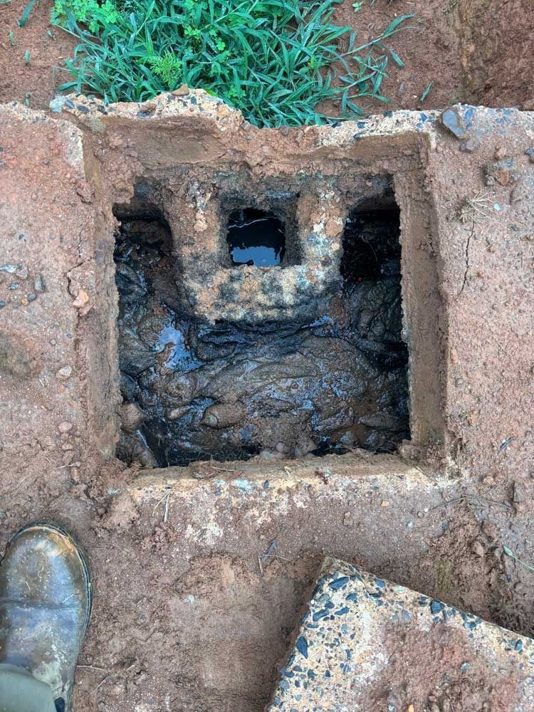 Open, dirt-surrounded drain with dark liquid inside. Soil and a boot are visible nearby.