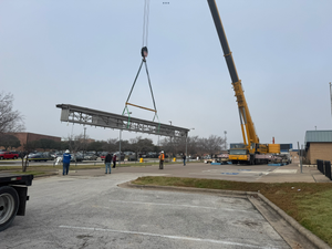 A crane lifting a long metal structure in a parking lot. Workers watch.