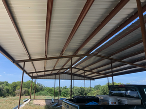 Metal roof shelter over a truck, set in a field. Brown metal beams and supports. Sky and vegetation in the background.