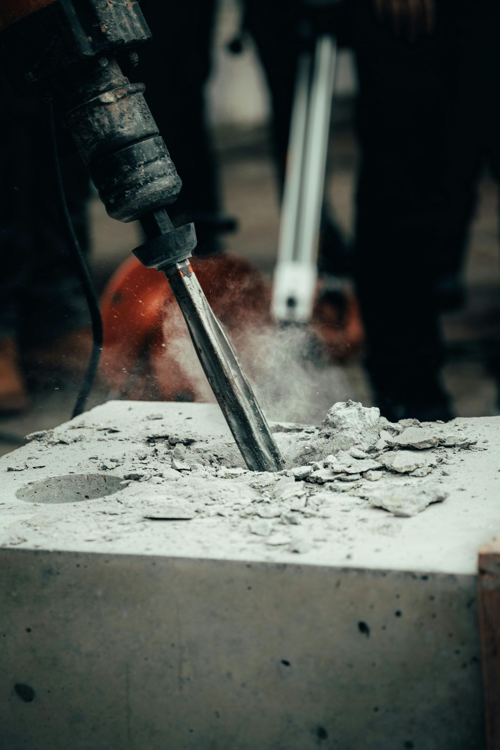 A jackhammer drills into a concrete block, creating dust; construction site.