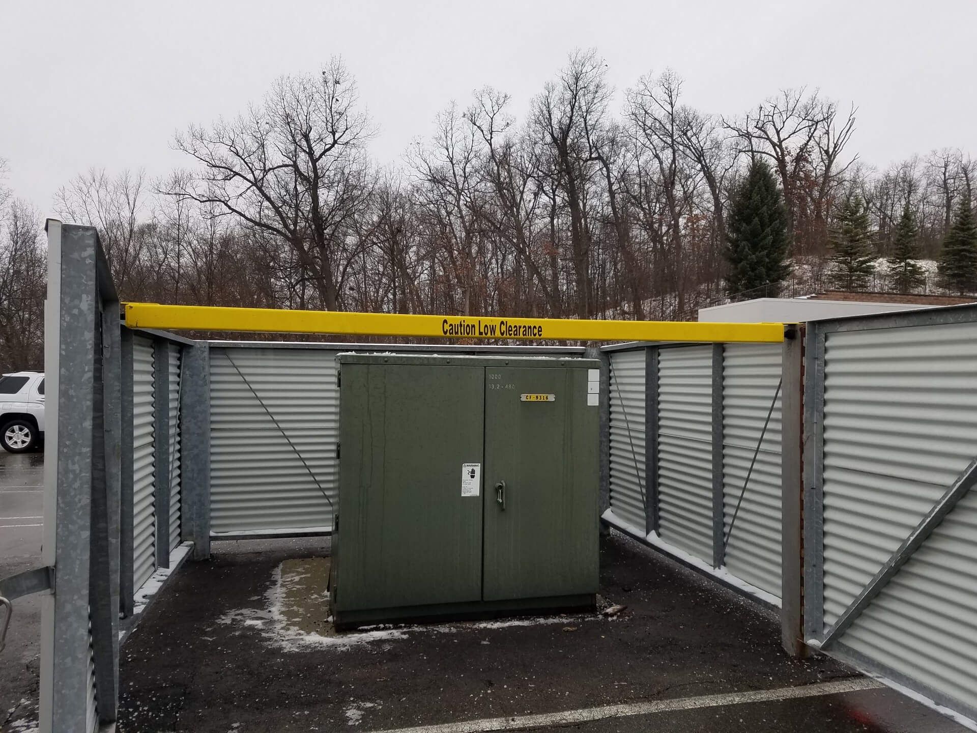 Metal-enclosed electrical transformer box with corrugated steel walls, yellow beam above, outdoor parking setting.