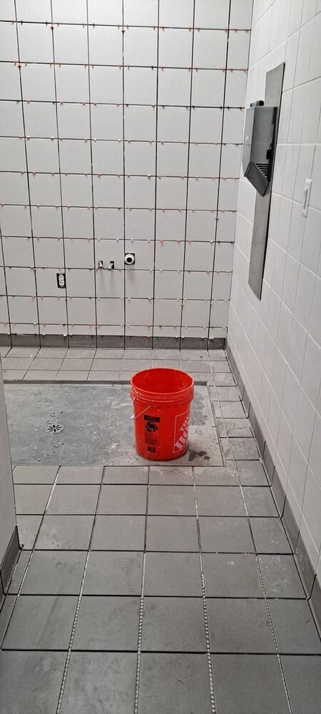 A bathroom under construction with white tiled walls, gray tiled floor, and a red bucket in the center.