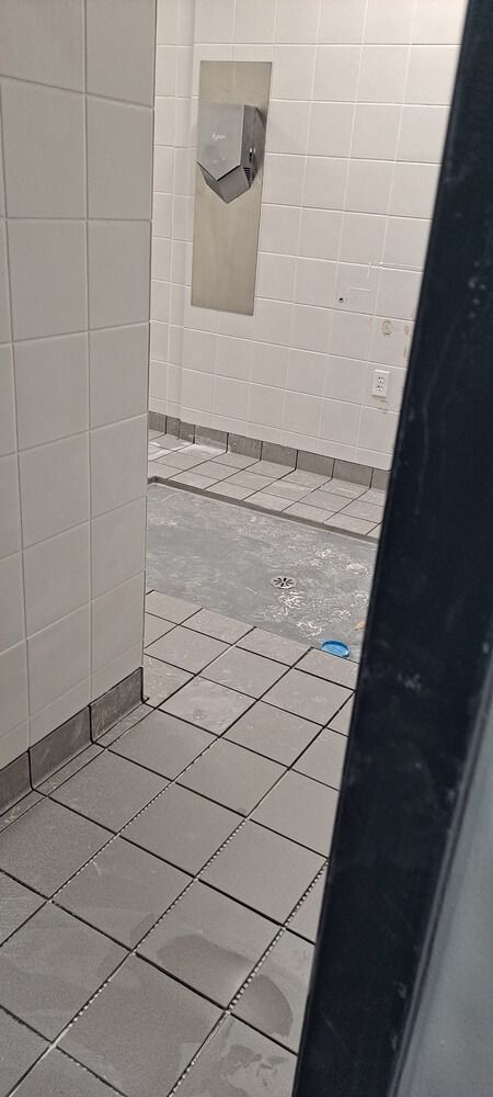 Interior view of a public restroom. White tiled walls and floors, hand dryer on the wall, and a partially obscured dark frame.