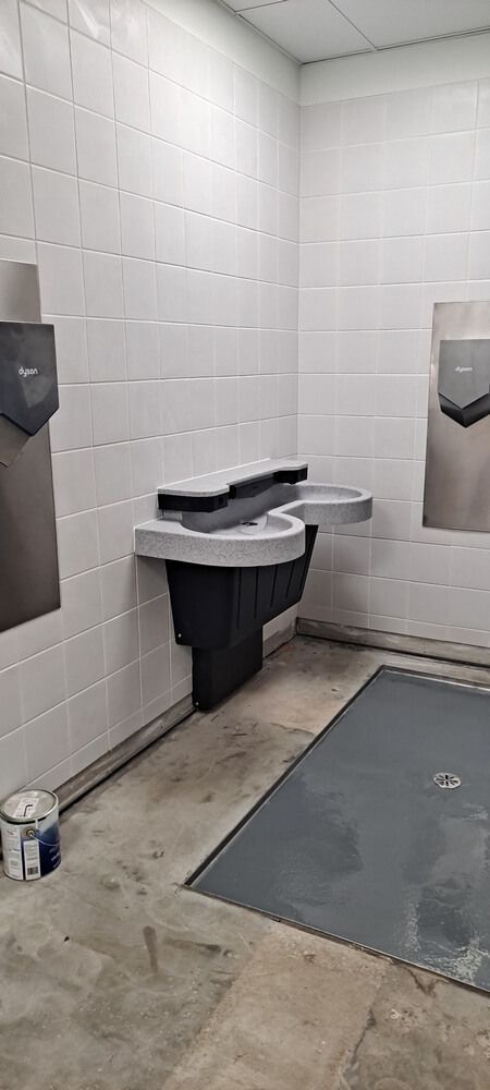 Public restroom with wall-mounted sink, hand dryers, and a paint can. Gray, white, and stainless steel colors.