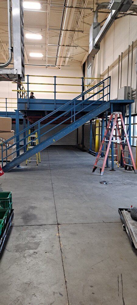 Warehouse interior with stairs leading to a platform. Ladders, equipment, and concrete floors are visible.