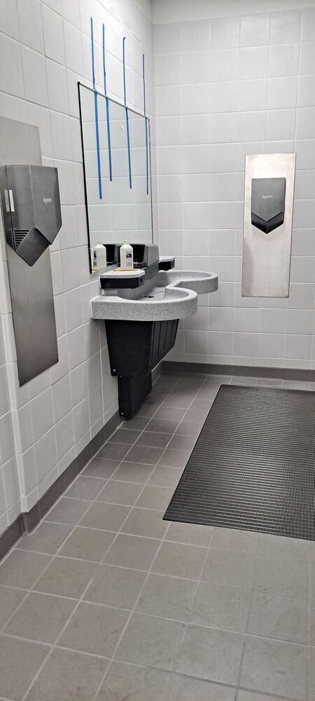 Public restroom with sinks, mirror, hand dryers, and a floor mat. Gray and white color scheme.