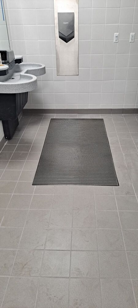 A restroom with gray tile, a mat, sinks, and a hand dryer on the wall.