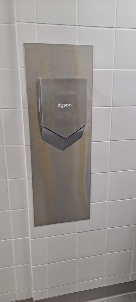 A Dyson hand dryer mounted on a stainless steel plate in a public restroom with white tiled walls.