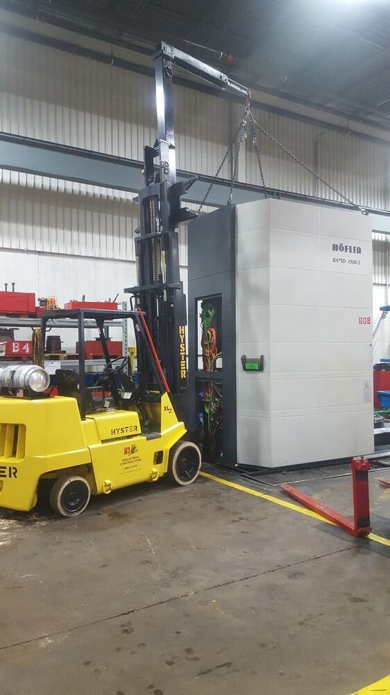 Yellow forklift lifting a large gray industrial machine in a factory.