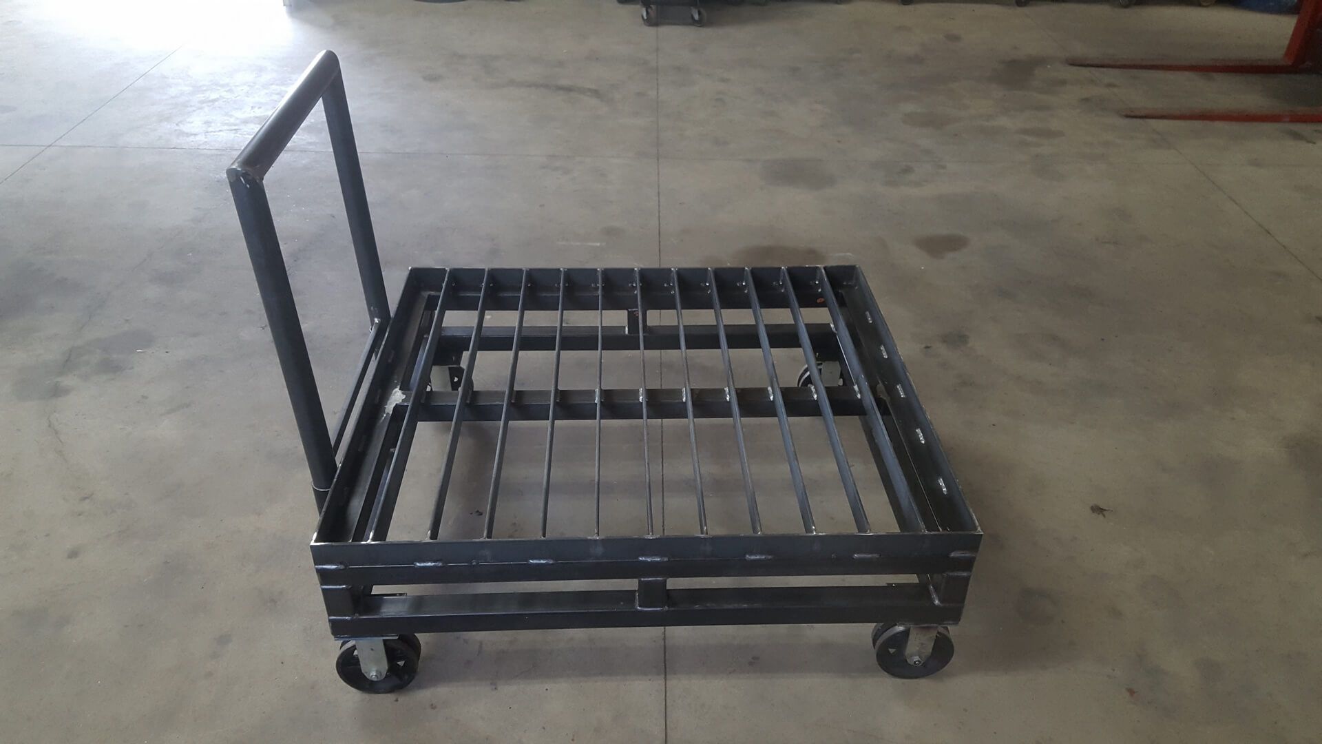 Black metal cart with a grated platform and a handle, on wheels.