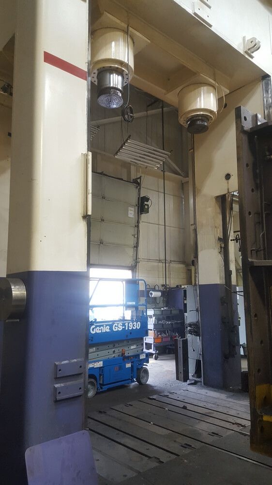 A large industrial stamping press with overhead lights, blue scissor lift, and open bay.