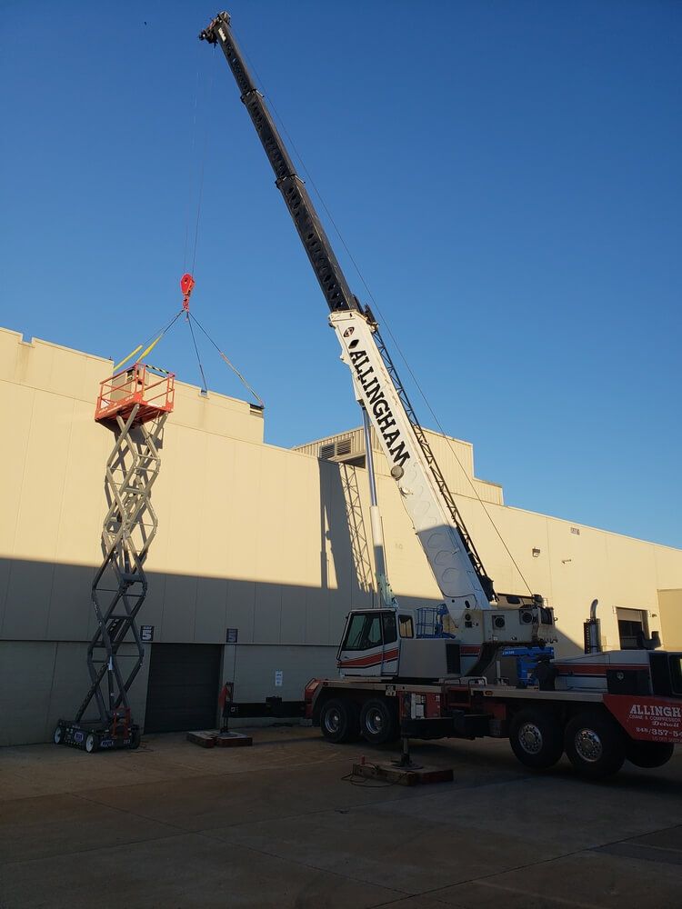 A large crane lifting a scissor lift with a load, outdoors next to a building.