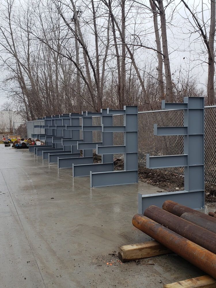 Steel support structures along a chain-link fence, with pipes on the ground.