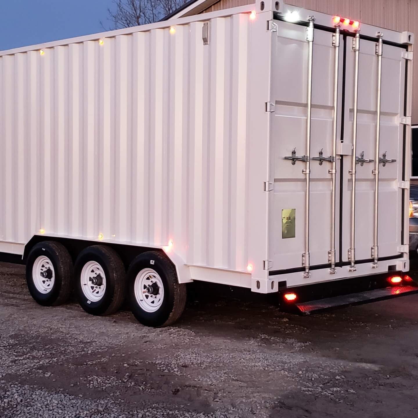 White container on a trailer with three wheels, illuminated with lights, against a dusky background.