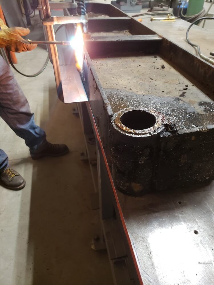 Person welding metal with a torch in a workshop. A large black machine part is visible.