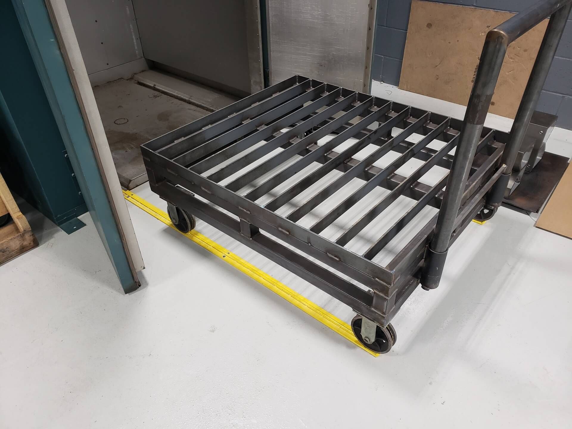 Metal cart with grated top and wheels, handle, near a yellow safety line and door.
