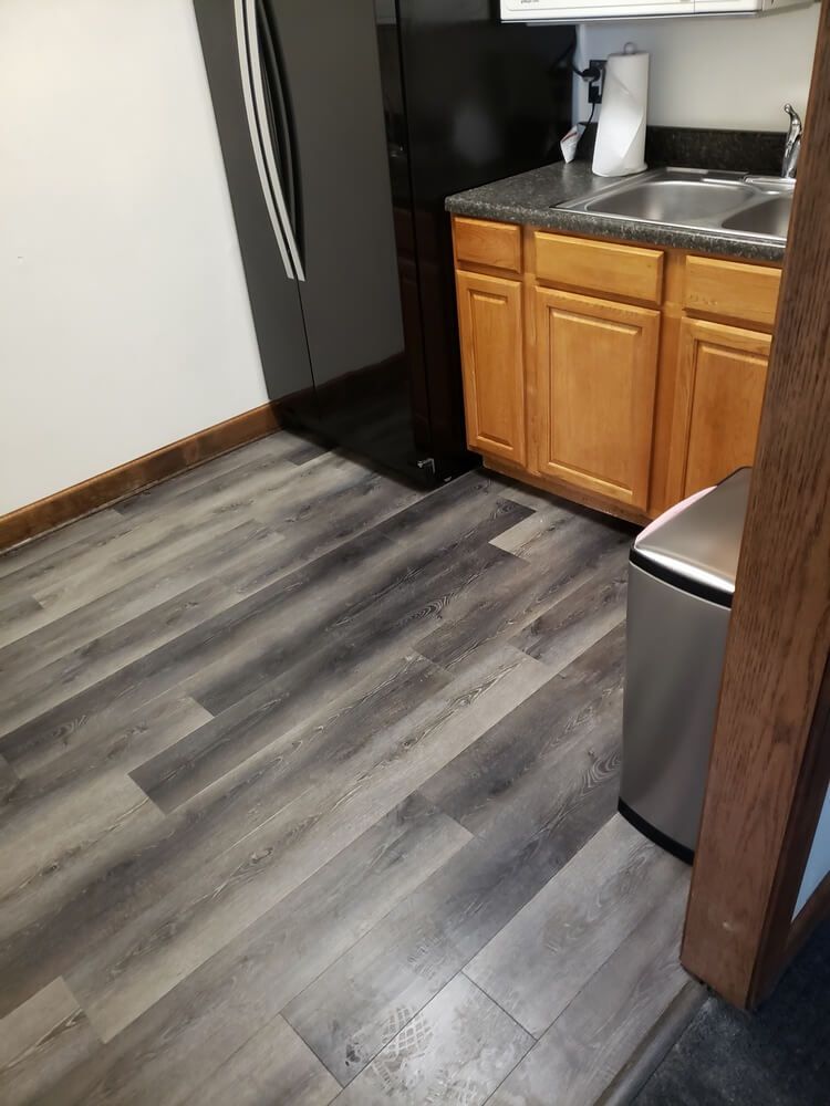 Kitchen with gray wood-look flooring, light wood cabinets, black refrigerator, and stainless steel trash can.