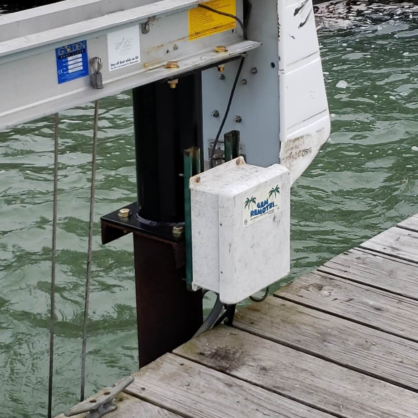 Boat lift mechanism attached to a wooden dock over water. White box, gray metal, and black pole.