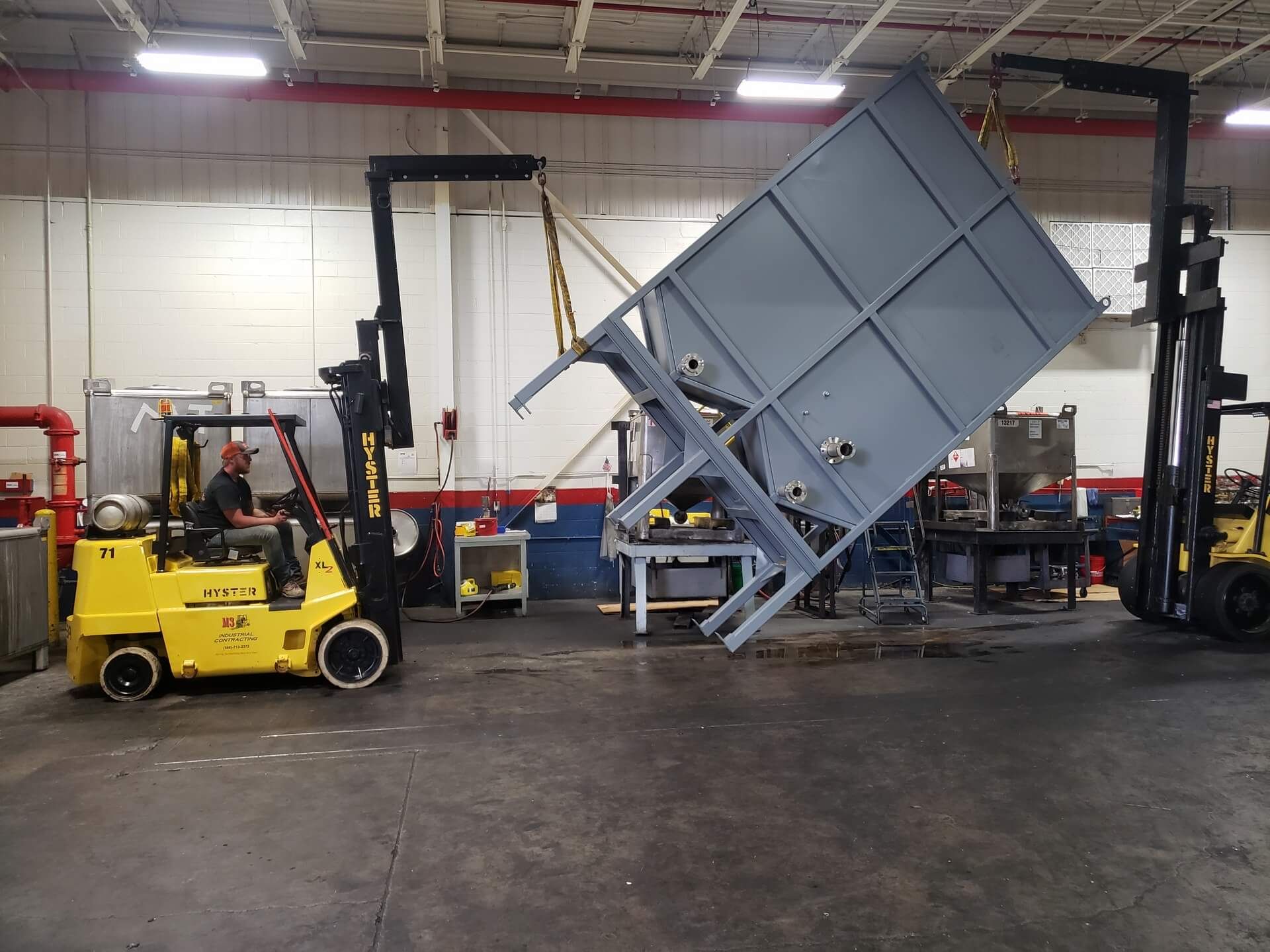 Two forklifts lifting a large, gray metal structure inside a warehouse. One forklift has an operator.