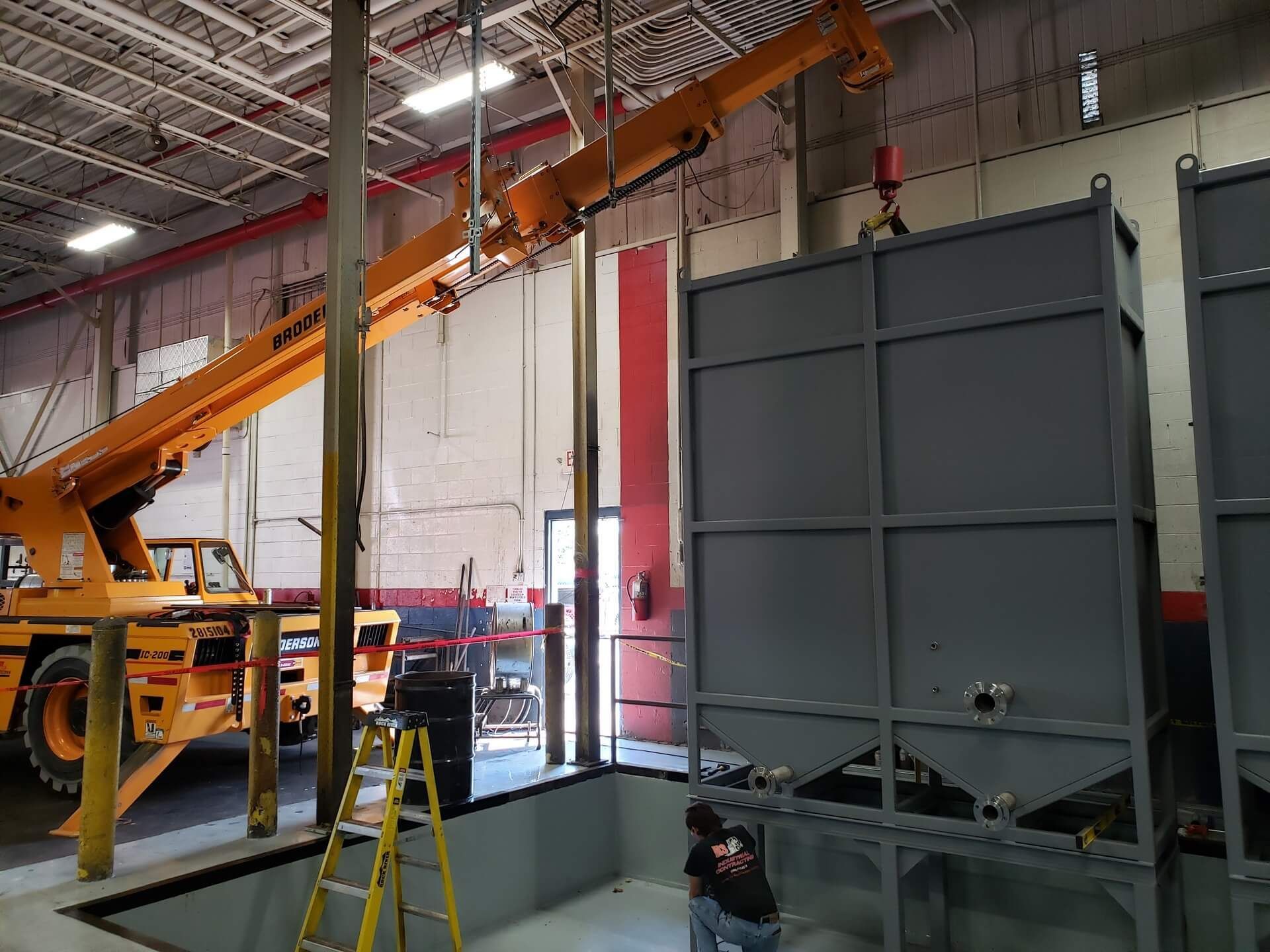 A crane lifting a large gray industrial tank inside a warehouse with a person working below.