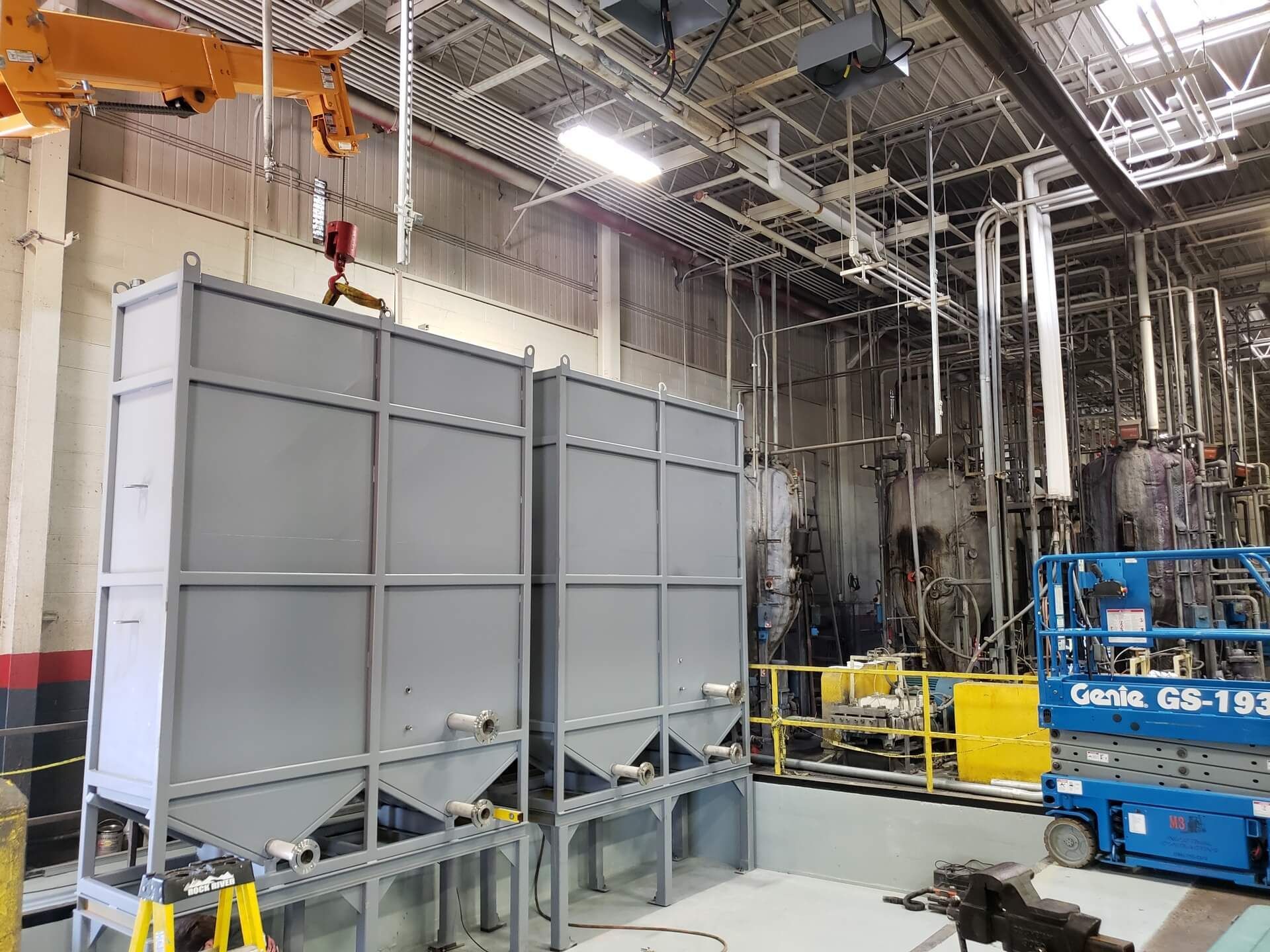 Two large gray industrial tanks in a factory setting with a crane overhead and a lift to the right.