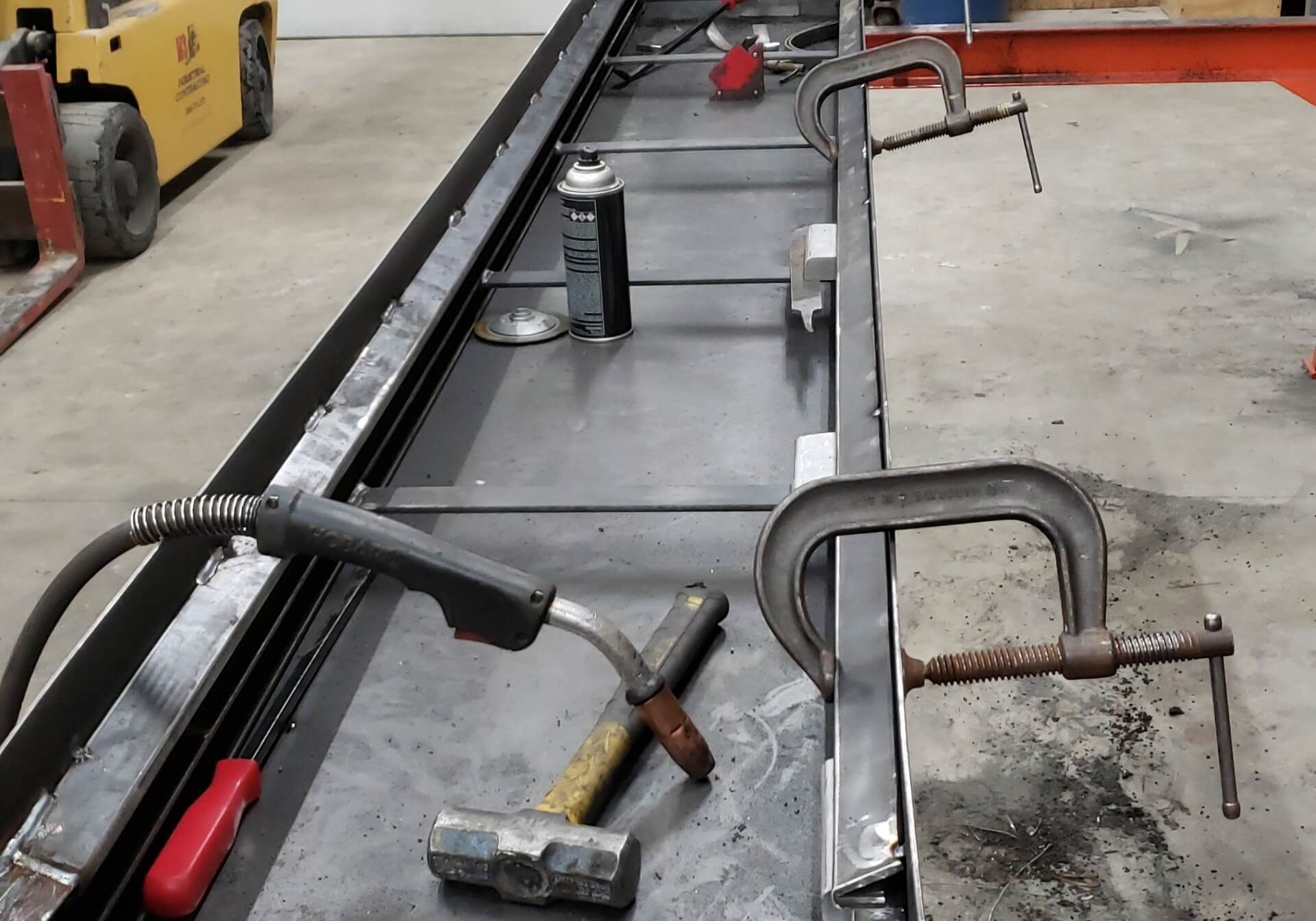 Welding setup on metal table: clamps, hammer, welding gun, spray can, and metal frame.