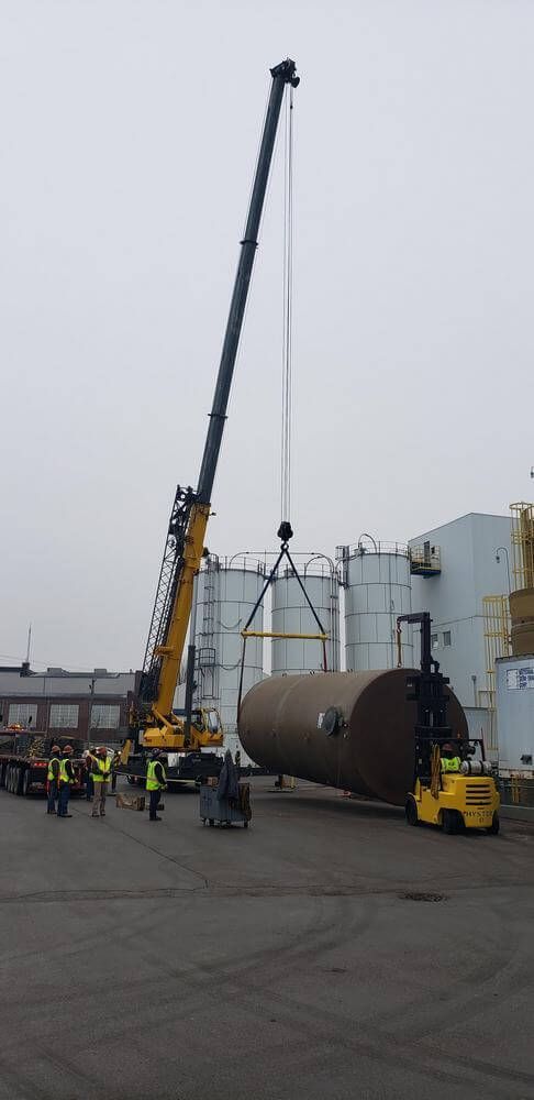A crane lifting a large cylindrical object with a forklift nearby; industrial setting.