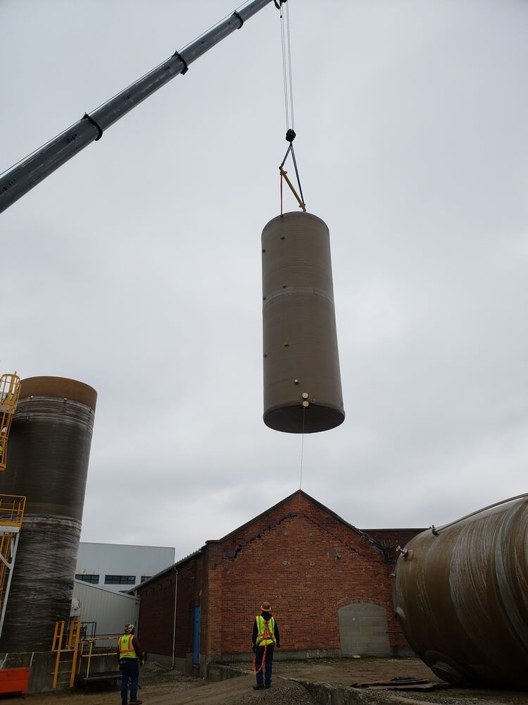 Crane lifting a large cylindrical tank near a brick building, with workers observing below.