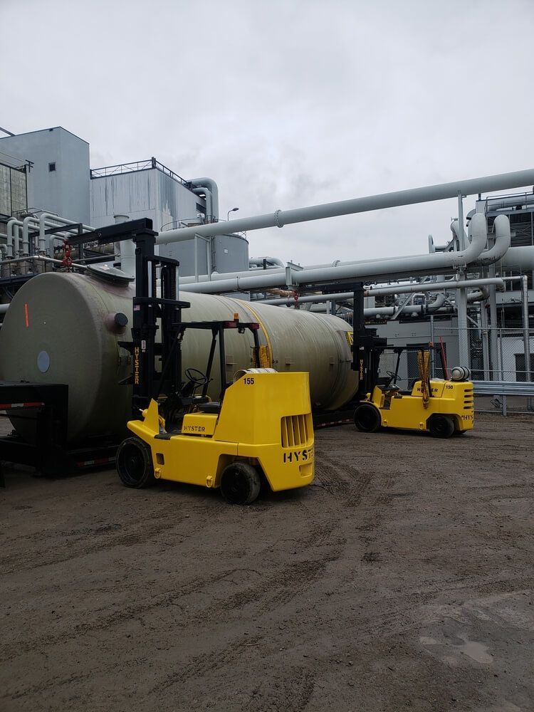 Two yellow forklifts near a large cylindrical tank at an industrial facility with pipes.
