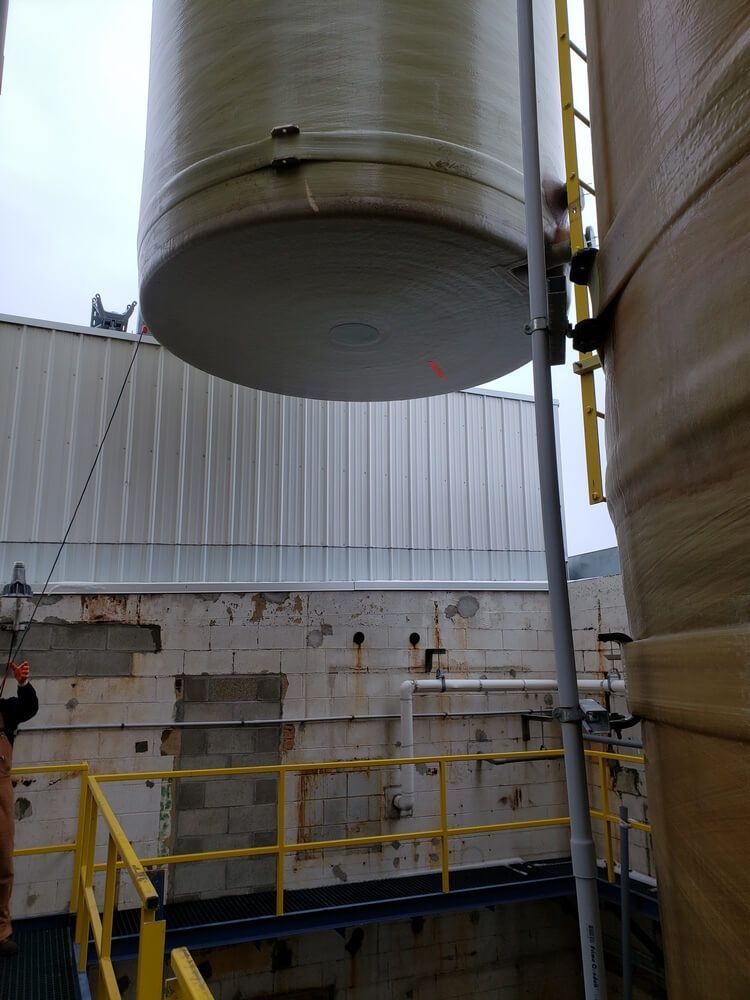 Large cylindrical tank being lifted above a building. Yellow safety railings and ladder visible.
