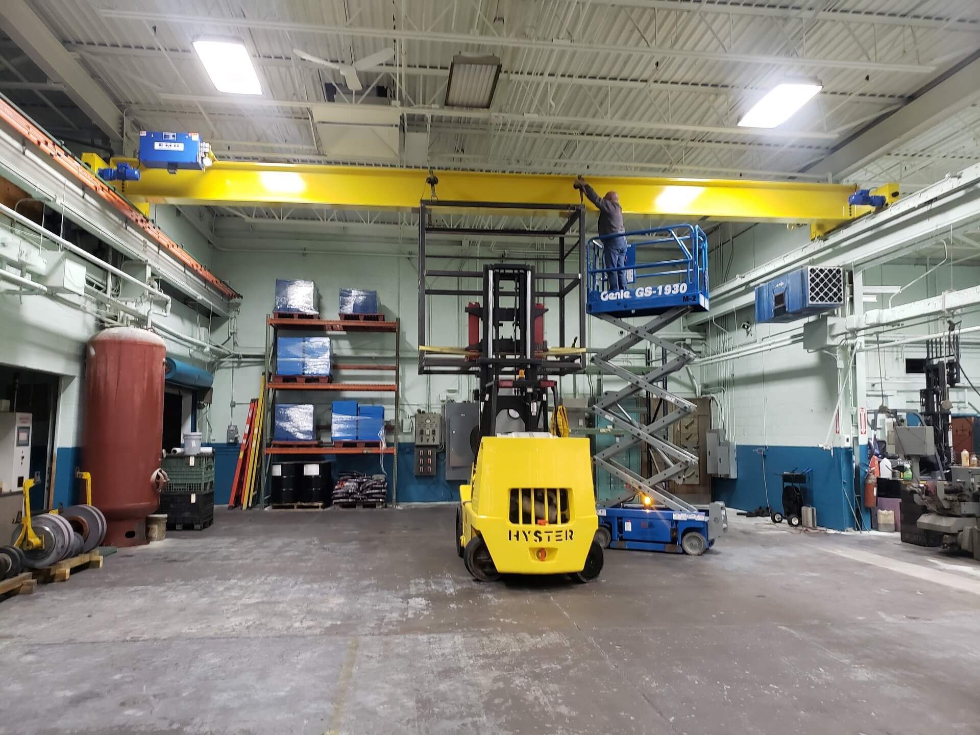Yellow forklift and scissor lift in a warehouse; worker on lift near yellow overhead crane.
