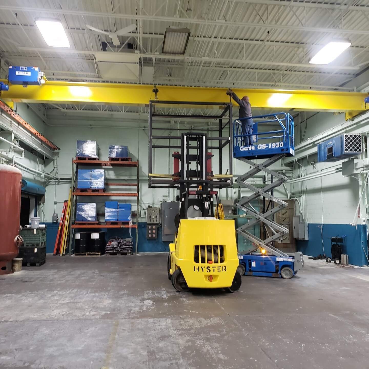 A person on a lift works on a yellow overhead crane in a factory. A forklift and shelving units are visible.