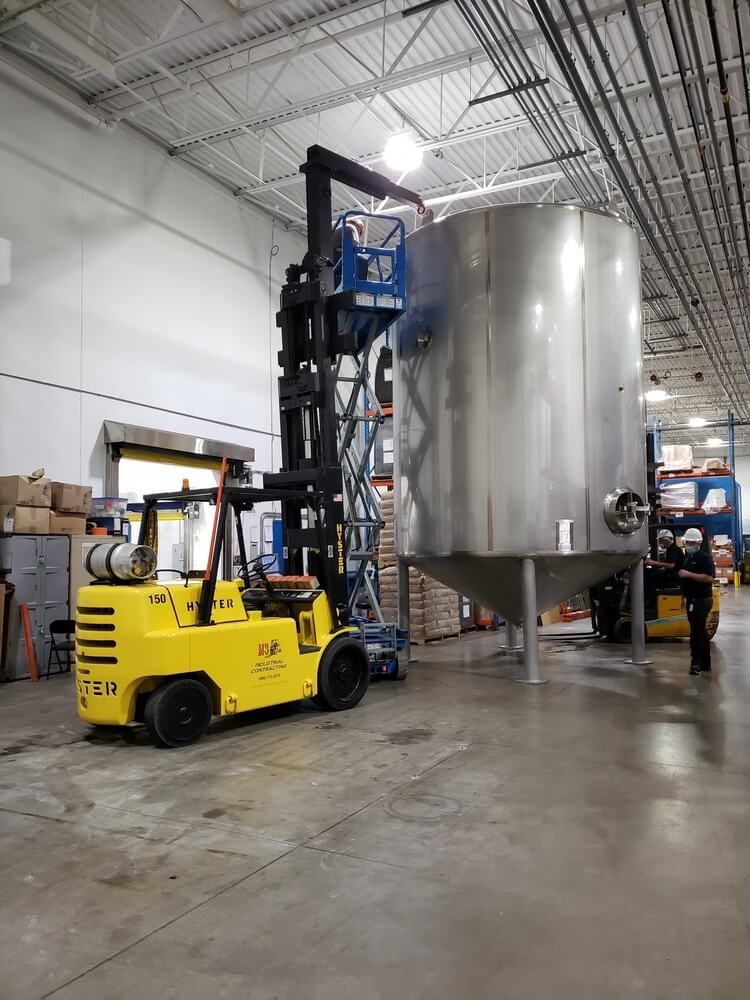 Yellow forklift with aerial lift basket near large silver industrial tank inside a warehouse.