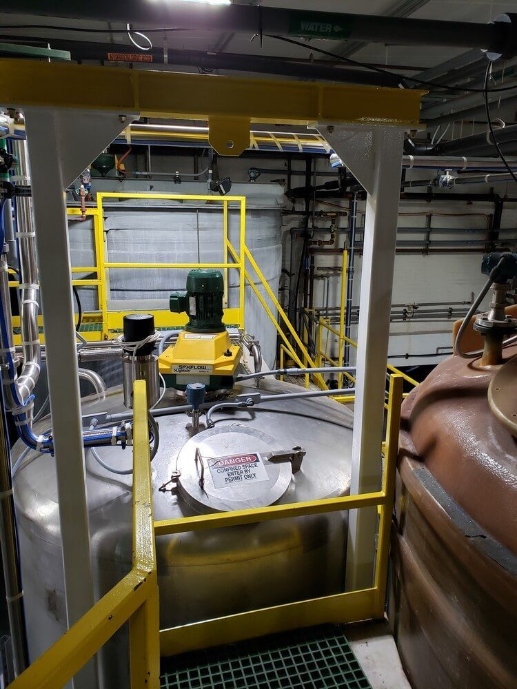 Stainless steel tank with motor in industrial setting, yellow and white safety structures.