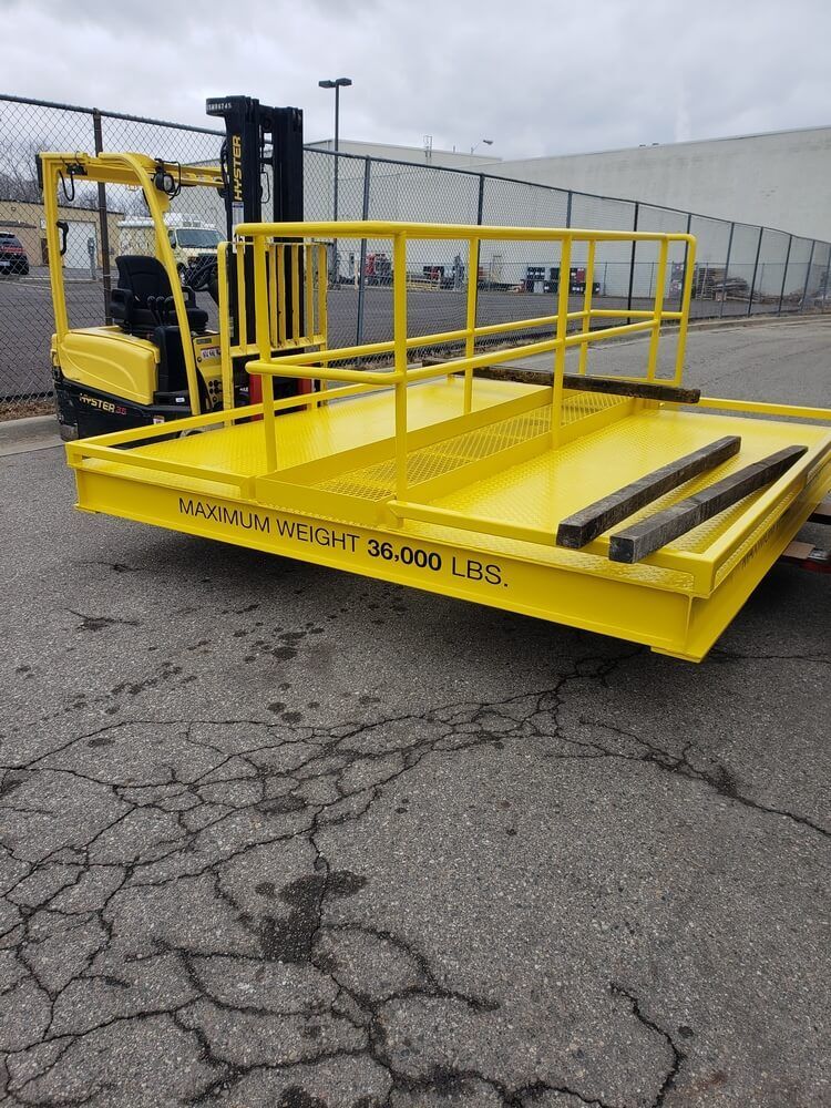 Yellow forklift platform with railing, parked on cracked pavement. The platform reads 