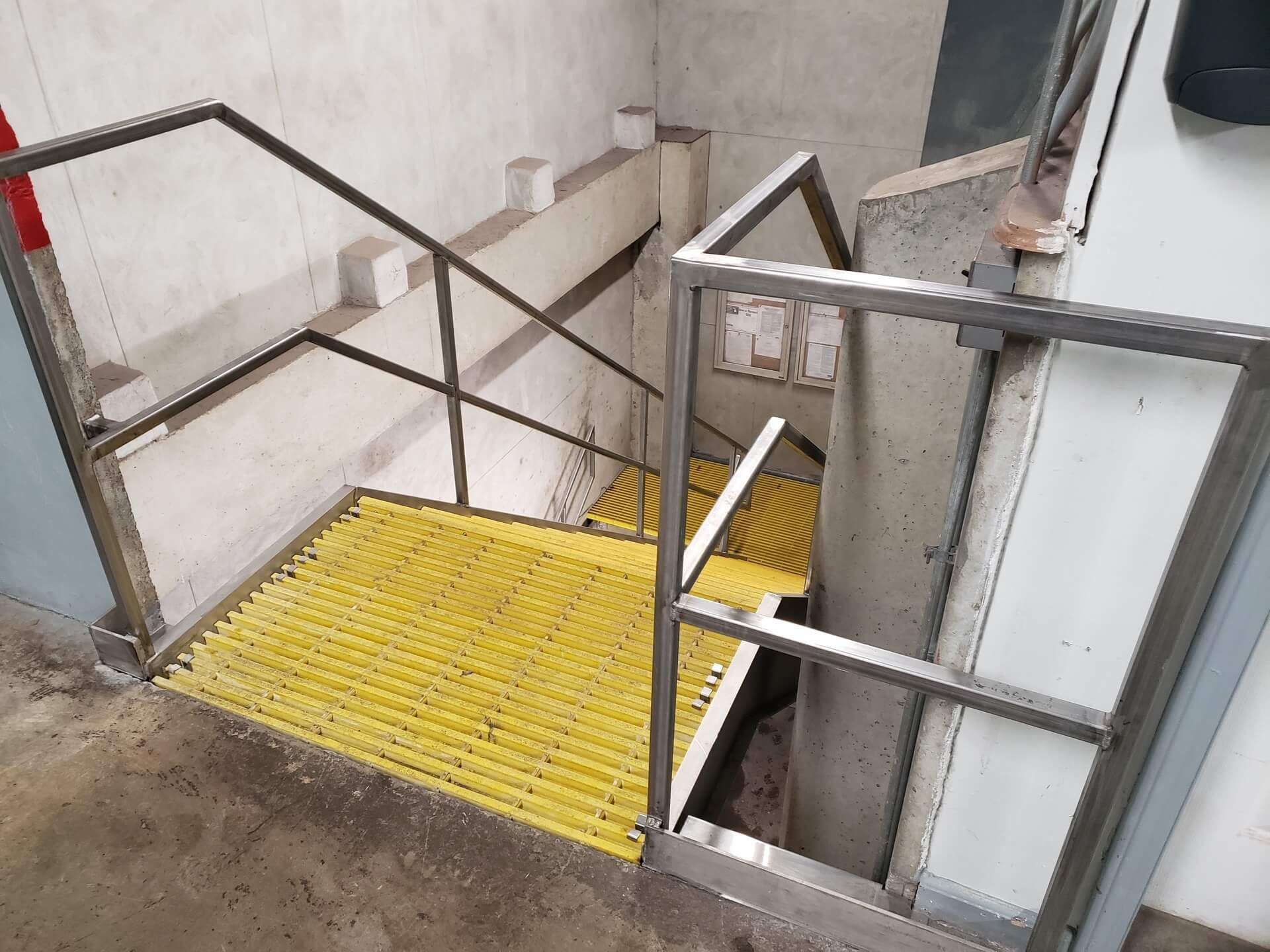 Staircase with yellow non-slip treads descends between concrete walls and metal railings.