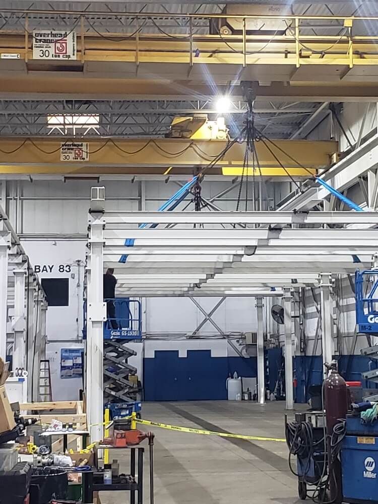 Man on lift works on metal structure under overhead crane in industrial setting.